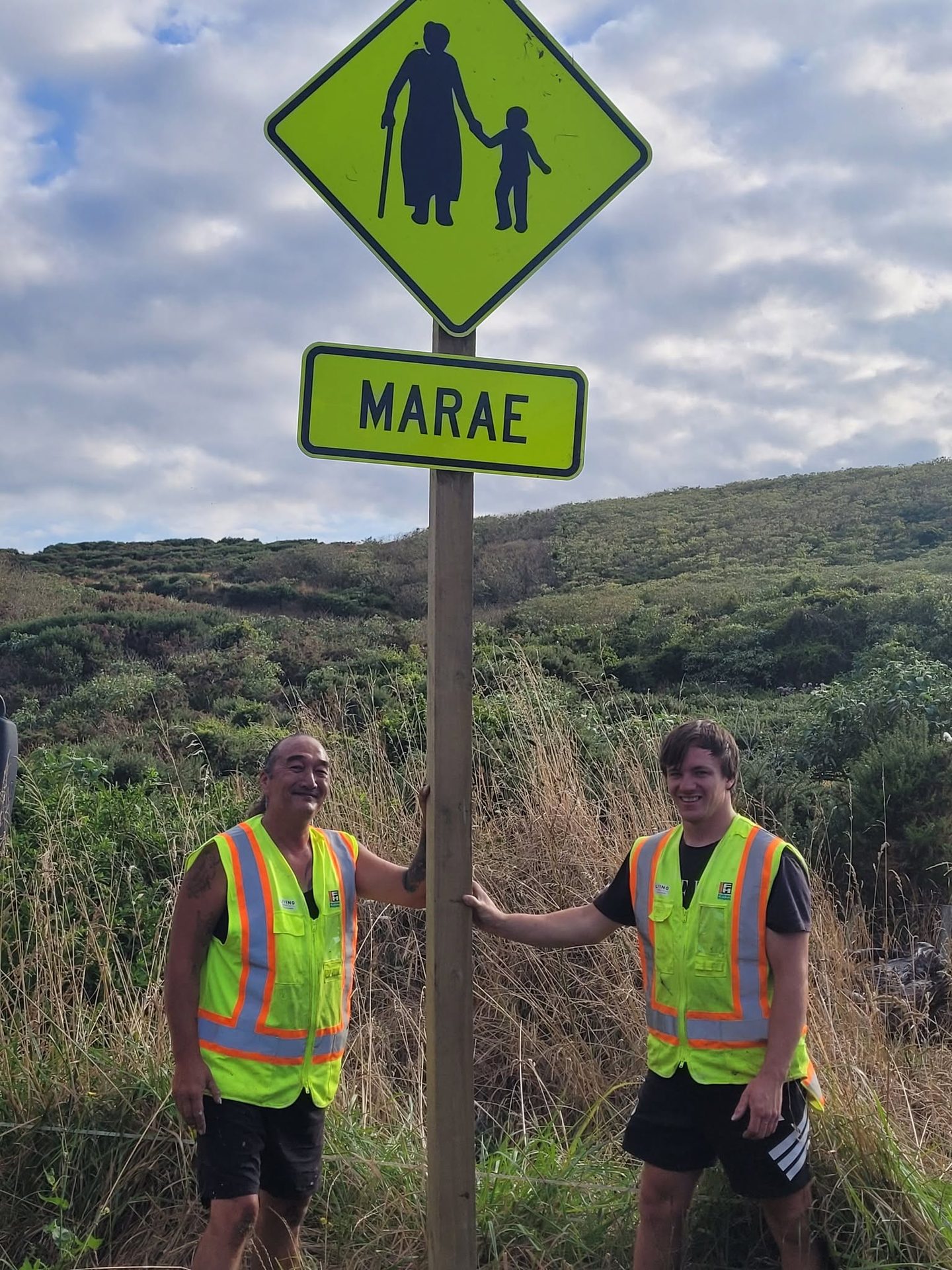 Two men smiling beside the marae sign